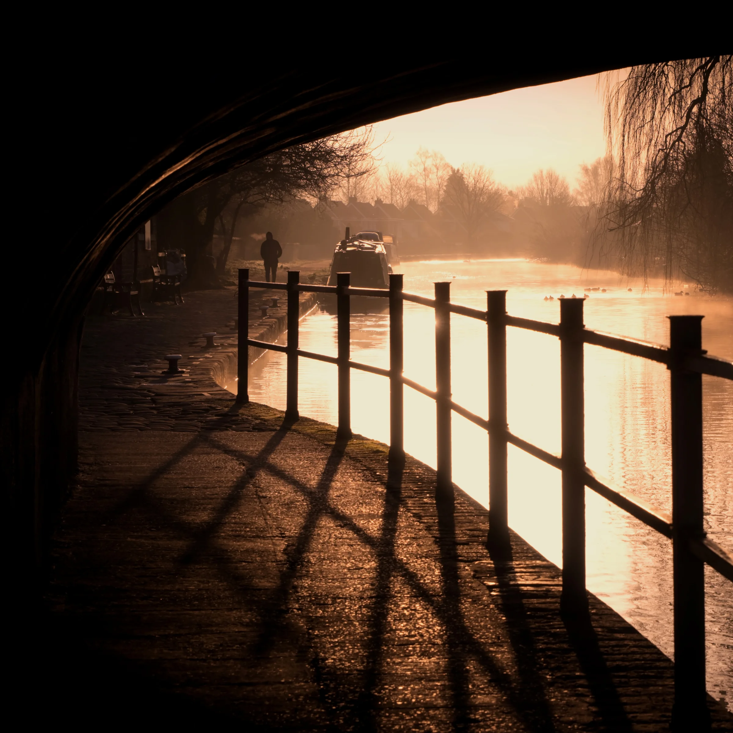 There's a strong chance this image contains the following subjects: sky,  fog,  light,  landscape,  sunset,  fall,  dawn,  dark,  lancashire,  sun,  bridge,  burscough,  ormskirk,  canal,  winter. The photographer has provided the following description: Near where I live, the morning fog was floating along the canal. I decided to duck under a bridge in order to frame the shot and give a little foreground interest. And then Mr Photobomber popped into shot and made it even better. I used to remove people from my images, but sometimes, it just works great! Photographer_name: Chris Michael. Title: Foggy Canal in Burscough. The photographer has provided the following description: Near where I live, the morning fog was floating along the canal. I decided to duck under a bridge in order to frame the shot and give a little foreground interest. And then Mr Photobomber popped into shot and made it even better. I used to remove people from my images, but sometimes, it just works great! Make: fujifilm. Model: X-T5. Technical: XF16-80mm lens. No filters. No colour grading. . Published: 2025-09-07 17:11:36
