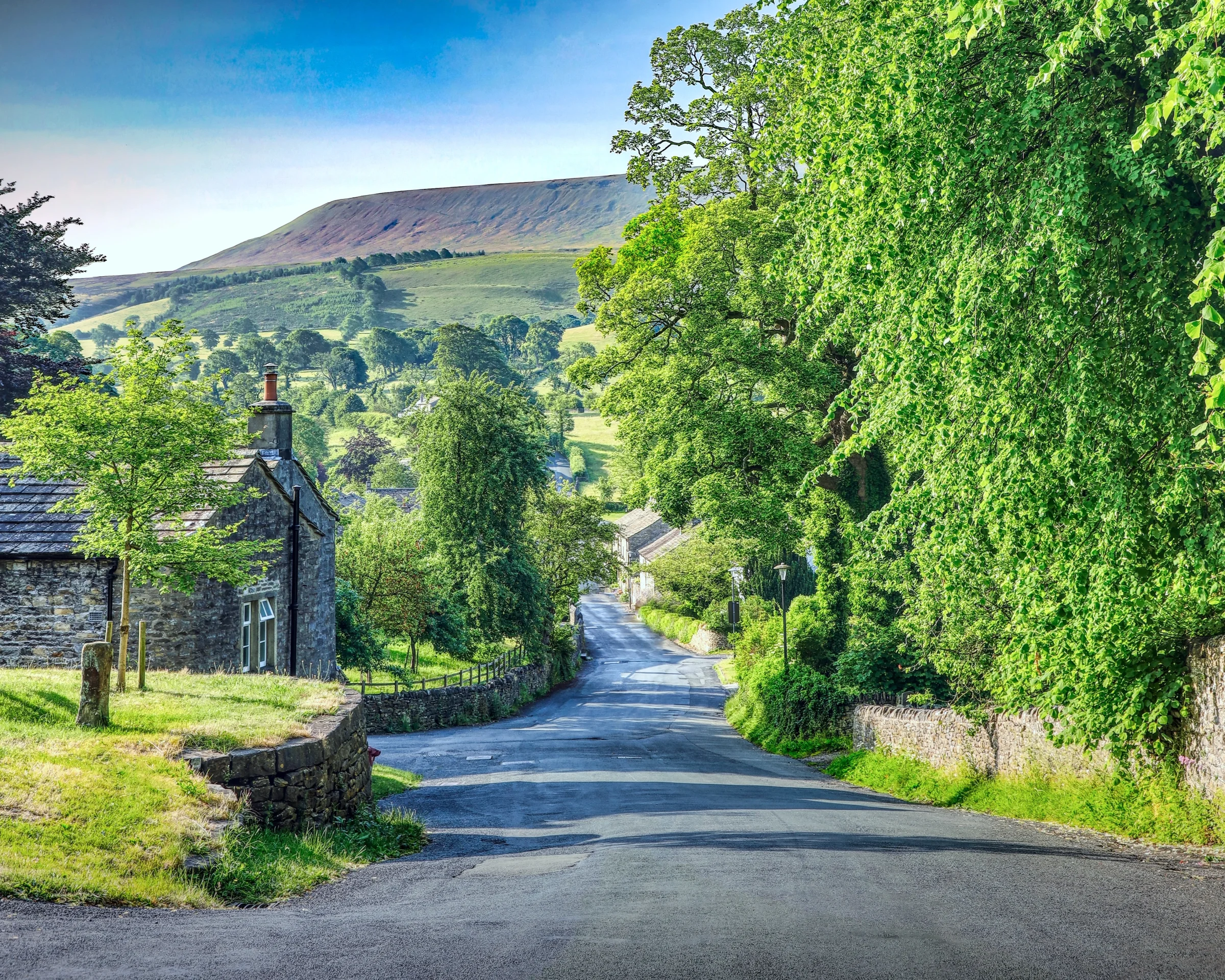 There's a strong chance this image contains the following subjects: tree,  landscape,  travel,  nature,  wood,  summer,  road,  lancashire,  rural,  countryside,  scenic. The photographer has provided the following description: This is the sleepy village of Downham in Lancashire, with Pendle Hill seen in the distance. This is a HDR bracketed shot. Photographer_name: Chris Michael. Title: Downham. The photographer has provided the following description: This is the sleepy village of Downham in Lancashire, with Pendle Hill seen in the distance. This is a HDR bracketed shot. Make: fujifilm. Model: X-T5. Technical: 16-80mm lens. Published: 2025-09-06 18:39:54
