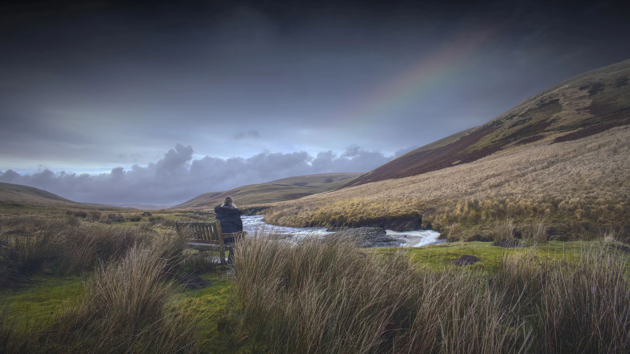 There's a strong chance this image contains the following subjects: sky,  water,  landscape,  lake,  nature. The photographer has provided the following description: Looks almost too 'fairytale' to be real..but it is. This is my wife, sat on a bench a couple of years ago looking into the distance at a rainbow. Taken in the Elan Valley around Powys, Wales. Photographer_name: Chris Michael. Title: Daydreaming. The photographer has provided the following description: Looks almost too 'fairytale' to be real..but it is. This is my wife, sat on a bench a couple of years ago looking into the distance at a rainbow. Taken in the Elan Valley around Powys, Wales. Make: canon. Model: EOS R. Technical: 16mm. Published: 2025-09-09 13:57:49