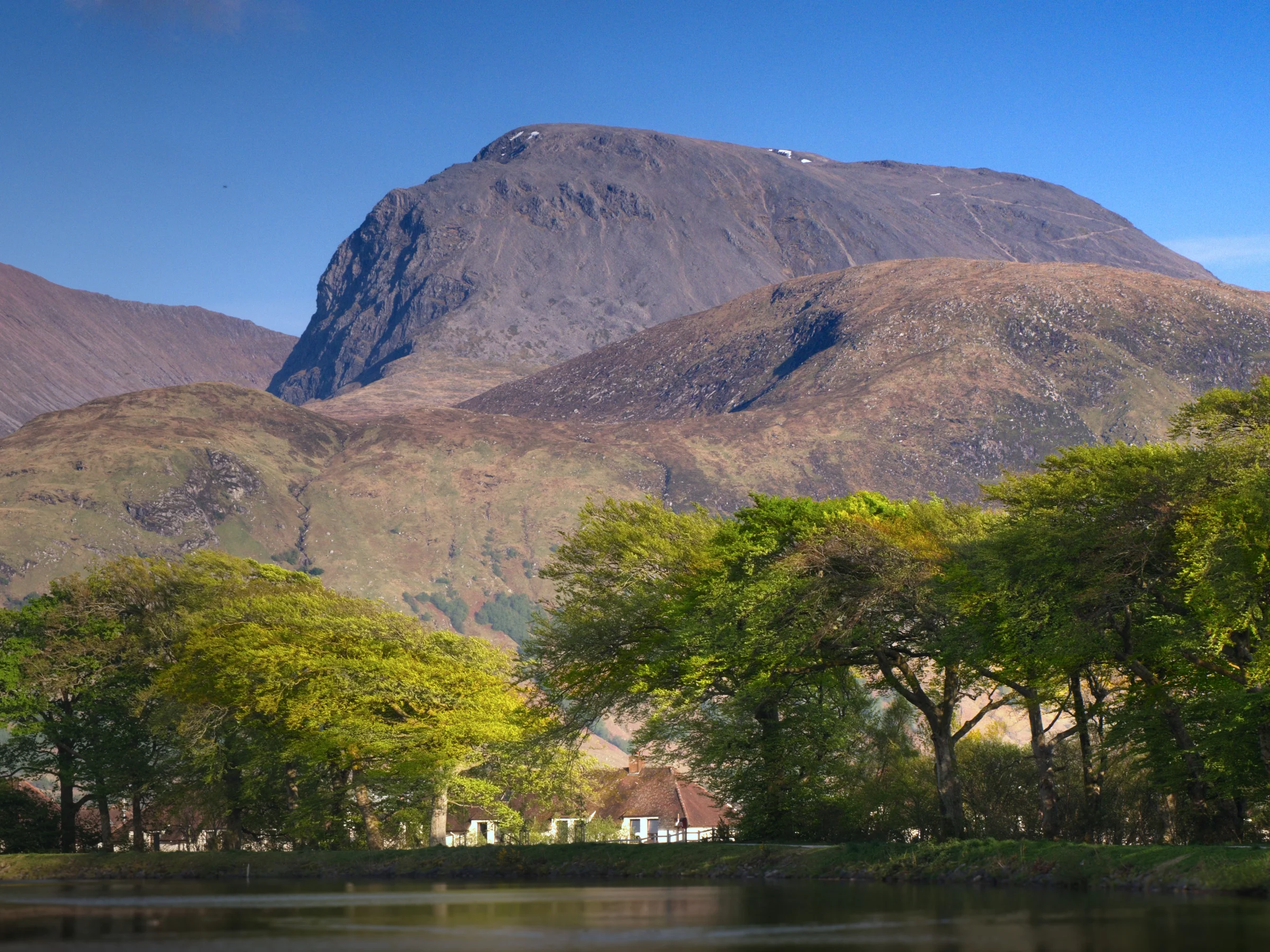 There's a strong chance this image contains the following subjects: tree,  sky,  water,  landscape,  lake,  travel,  nature,  river,  canal,  scotland,  corpach,  fortwilliam,  bennevis,  trees. The photographer has provided the following description: Scotland's highest mountain is Ben Nevis, seen here towering over Fort William. Taken from the side of the Caledonian Canal in Corpach. Photographer_name: Chris Michael. Title: Ben Nevis. The photographer has provided the following description: Scotland's highest mountain is Ben Nevis, seen here towering over Fort William. Taken from the side of the Caledonian Canal in Corpach. Make: fujifilm. Model: X-T5. Technical: 16-80mm at 80mm. Published: 2025-09-08 17:12:00