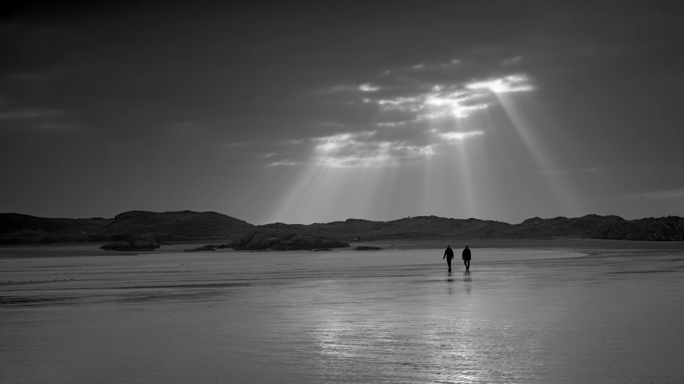 There's a strong chance this image contains the following subjects: monochrome,  water,  landscape,  sea,  beach,  reflection,  ocean,  seascape,  anglesey,  wales. The photographer has provided the following description: Whilst walking over to LLanddwyn Island hoping to catch the sunset, the clouds quickly moved in, and just as two people walked into the shot, the clouds opened up and streaked down a beam of light below.  Photographer_name: Chris Michael. Title: Heavens Above. The photographer has provided the following description: Whilst walking over to LLanddwyn Island hoping to catch the sunset, the clouds quickly moved in, and just as two people walked into the shot, the clouds opened up and streaked down a beam of light below.  Make: fujifilm. Model: X-T5. Technical: 16-80mm. Published: 2025-09-09 13:34:35