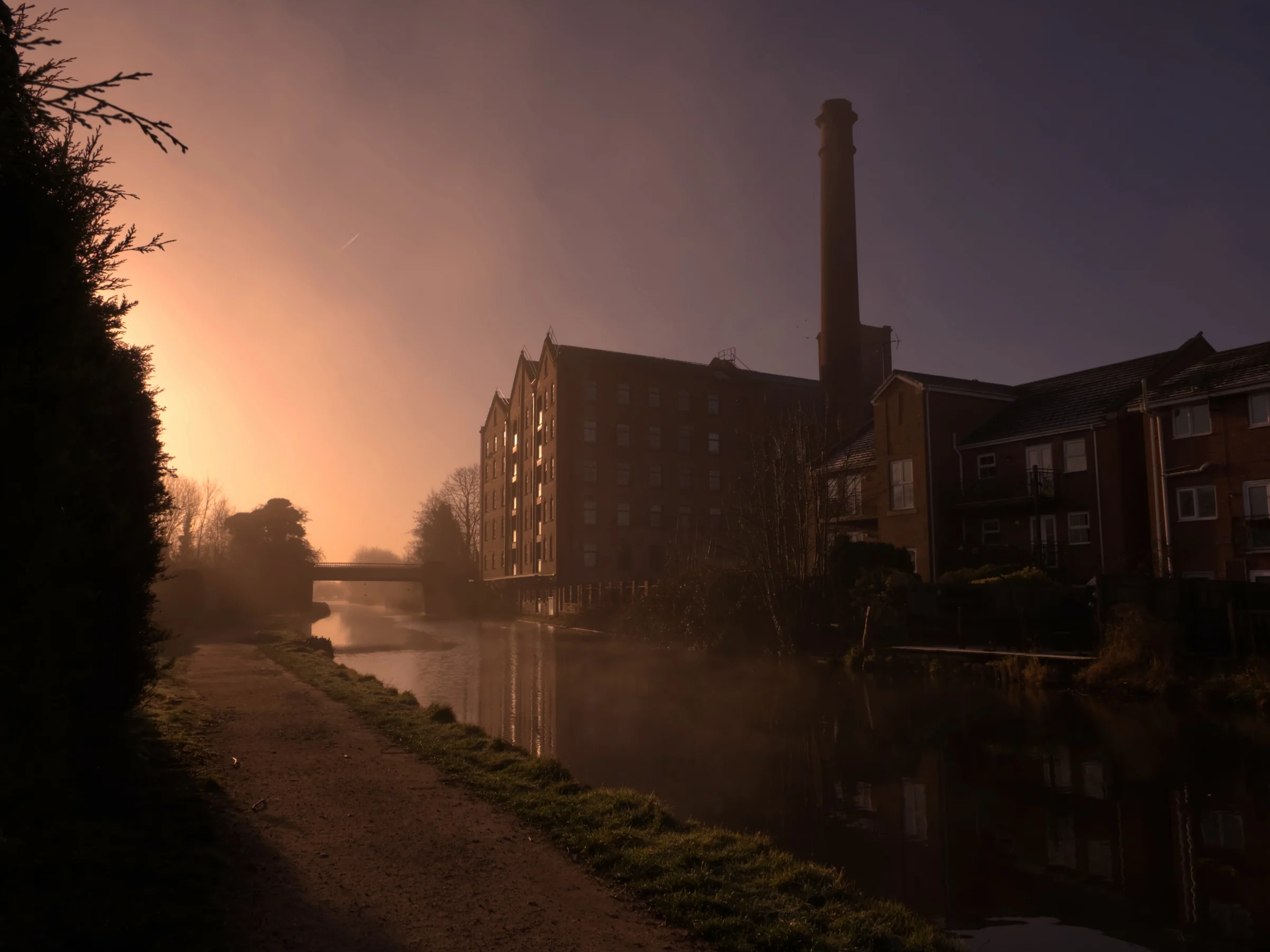 There's a strong chance this image contains the following subjects: street,  fog,  light,  water,  city,  sunset,  dawn,  lancashire,  bridge,  burscough,  ormskirk,  canal,  sunrise. The photographer has provided the following description: Just along the Leeds-Liverpool Canal, is Ainscough Mill at Burscough in Lancashire. This was taken around 7;15am during sunrise. Photographer_name: Chris Michael. Title: Foggy Morning in Burscough. The photographer has provided the following description: Just along the Leeds-Liverpool Canal, is Ainscough Mill at Burscough in Lancashire. This was taken around 7;15am during sunrise. Make: fujifilm. Model: X-T5. Technical: XF16-80mm lens. Published: 2025-09-06 18:10:27