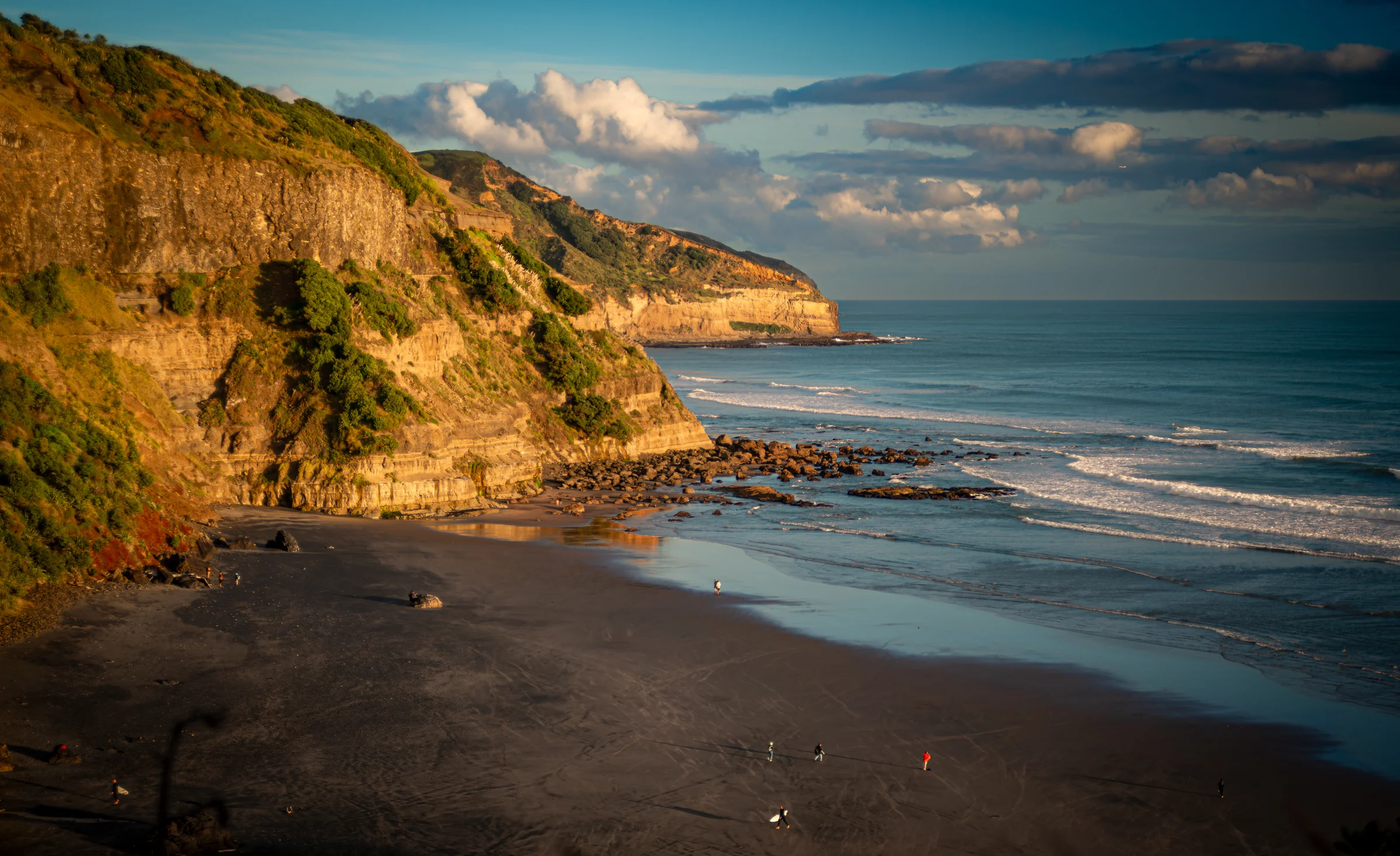 There's a strong chance this image contains the following subjects: landscape,  sea,  beach. The photographer has provided the following description: Muriwai Beach New Zealand Photographer_name: Robert A F van de Voort. Title: Black sand with surfers returning. The photographer has provided the following description: Muriwai Beach New Zealand Make: nikon. Model: D800. Technical: 210 - 70 mm Nikkor.. Published: 2026-06-03 04:16:14
